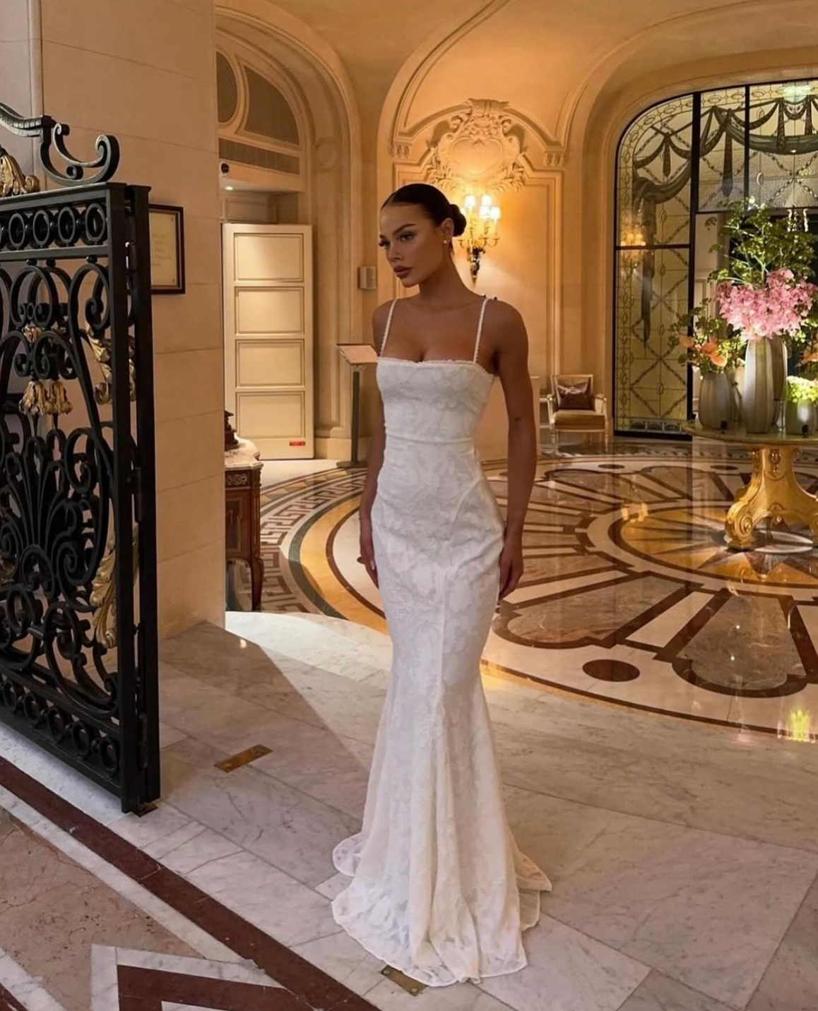 Woman in a white evening gown standing in an elegant interior setting with decorative elements.