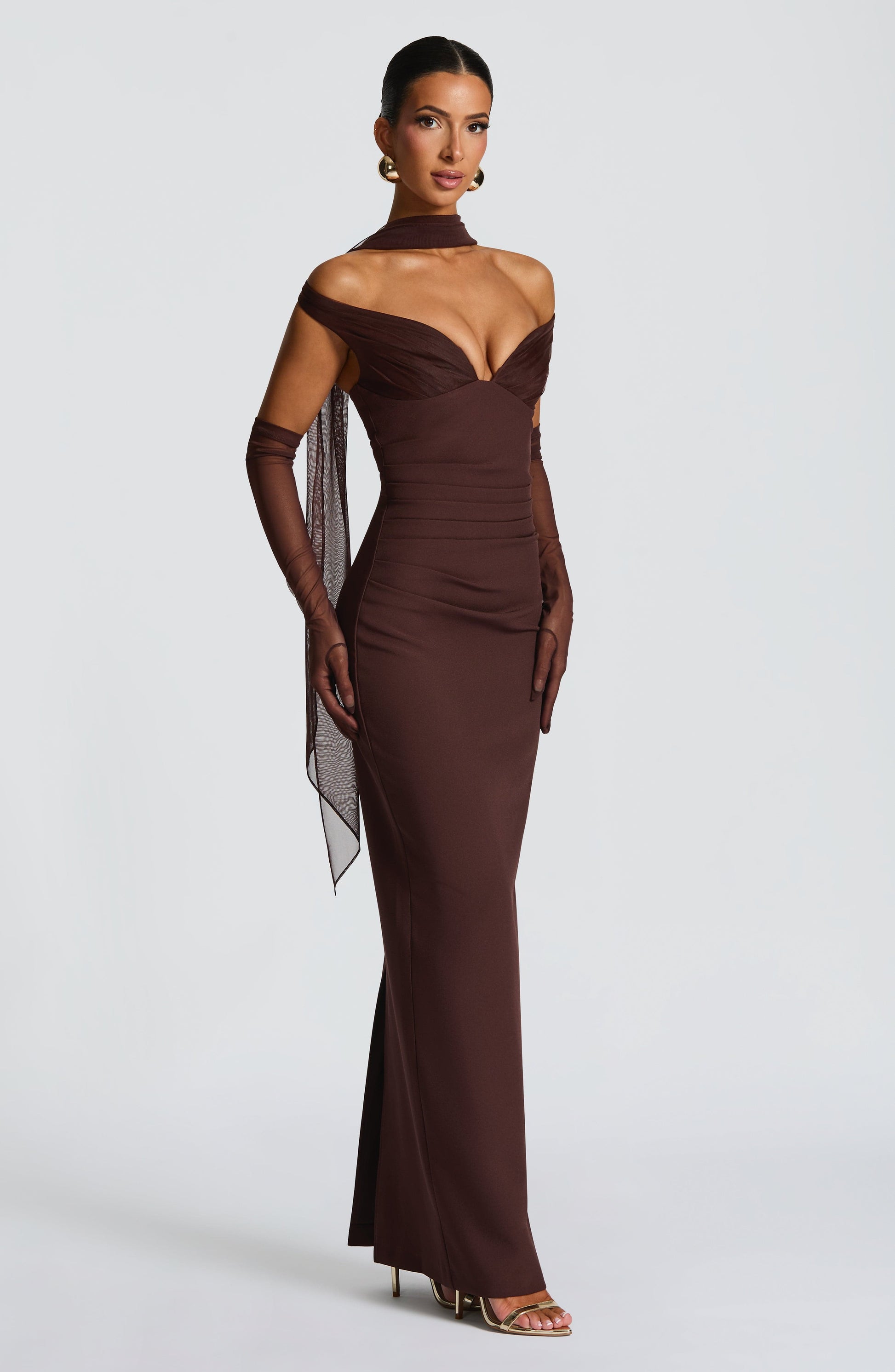 Woman wearing a brown off-shoulder evening gown with sheer sleeves on a white background