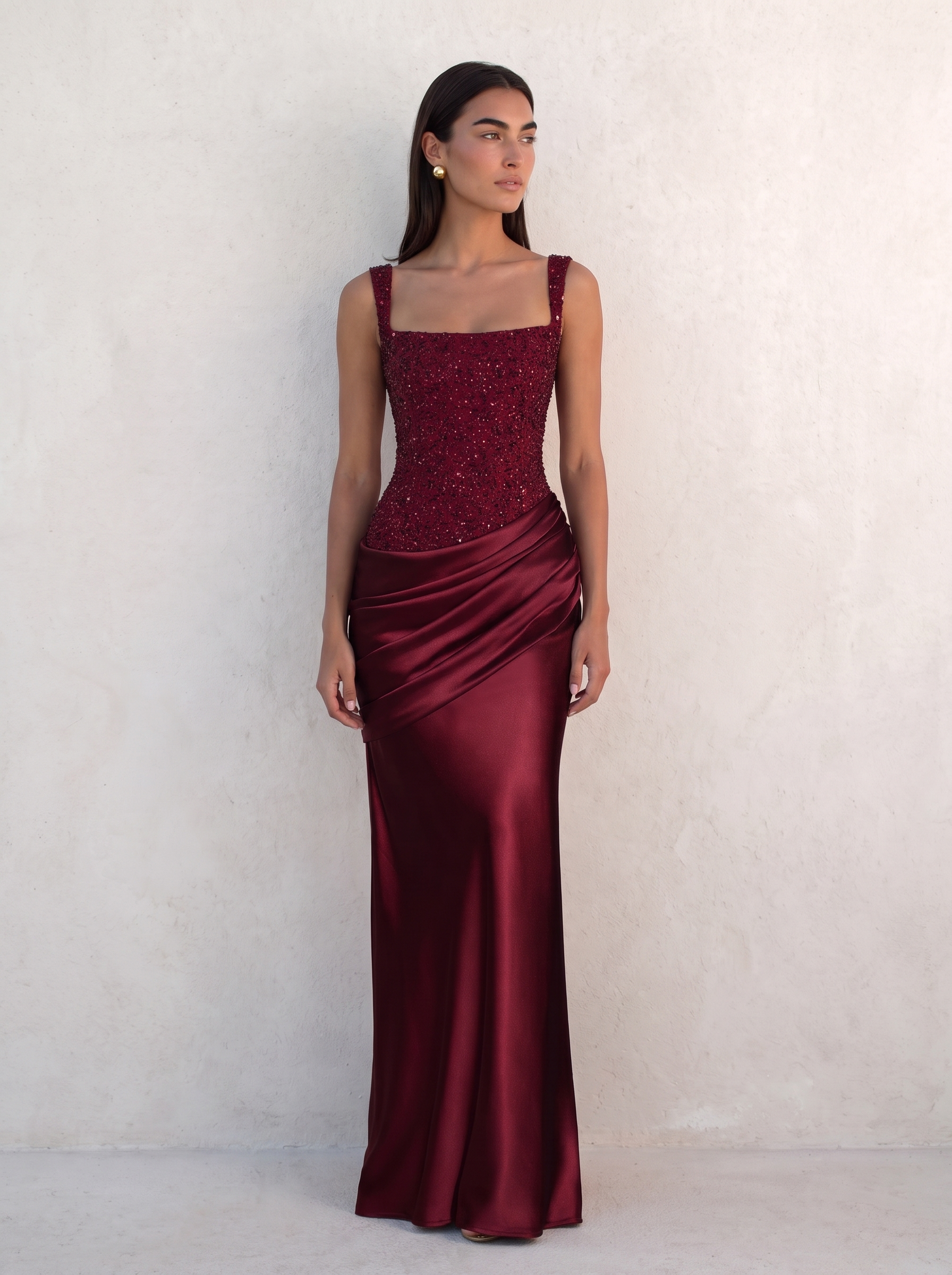 Model wearing the VESPERA BEADED GOWN in burgundy against a pale plaster wall — beaded corset bodice, square neckline and flared satin maxi skirt — Kalii