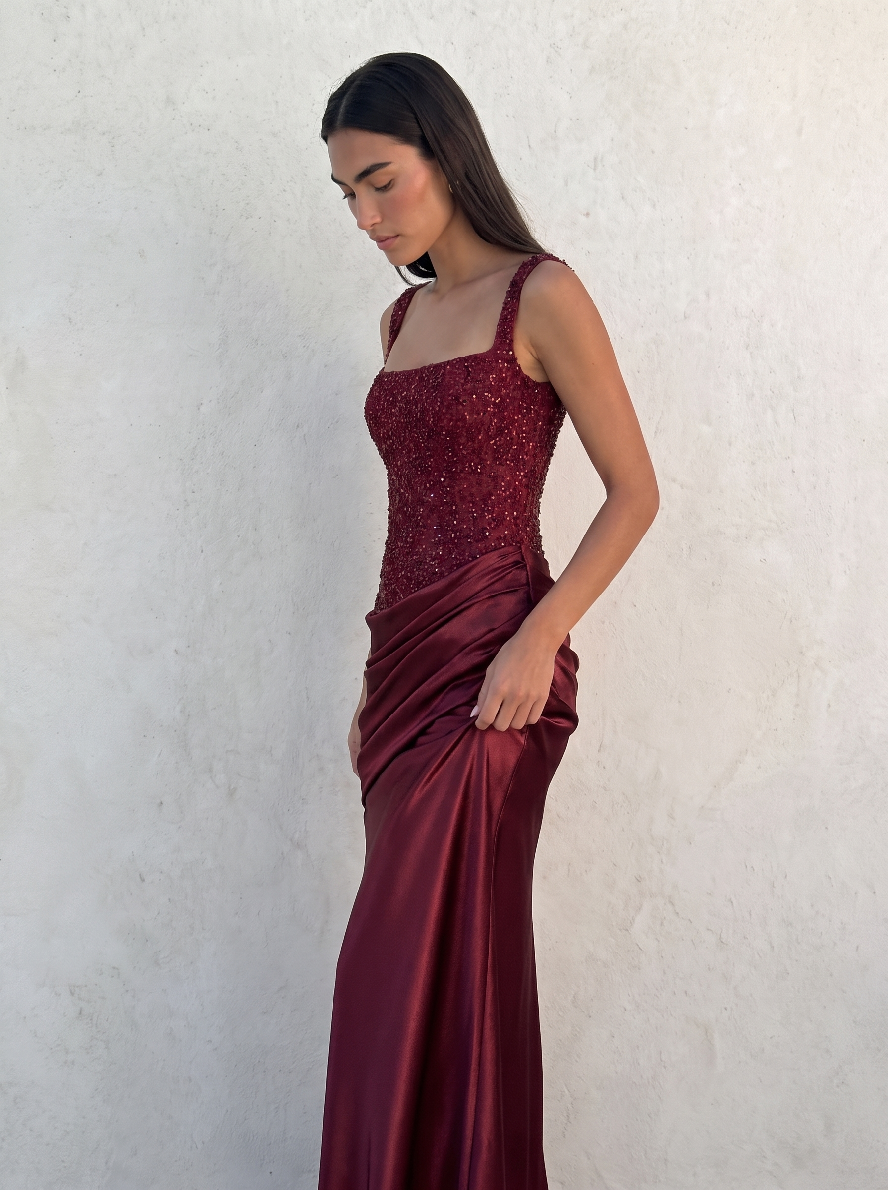 VESPERA BEADED GOWN in burgundy — three-quarter view showing the asymmetric pleated hip drape — Kalii